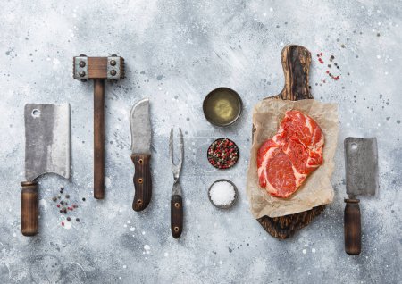 Photo for Rib eye steak with meat cleaver,fork,knife with wooden tenderizer and various herbs and oil with chopping board on light kitchen background.Top view. - Royalty Free Image