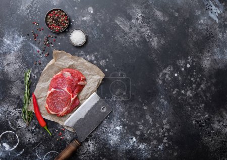 Foto de Trozo de filete de ternera roja cruda con cuchilla de acero, sal y pimienta sobre fondo de mesa oscuro con romero y pimienta roja. - Imagen libre de derechos