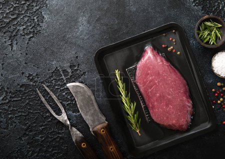 Photo for Beef raw fillet steak in vacuum tray with pepper,salt and rosemary on black background with barbeque fork and knife.Top view - Royalty Free Image
