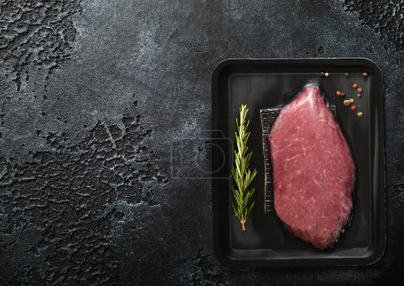 Téléchargez les photos : Steak de filet de boeuf cru frais scellé dans un bac à vide avec poivre et romarin sur fond de table de cuisine noire.Vue du dessus. - en image libre de droit