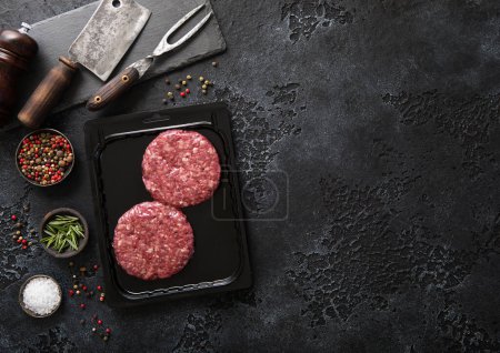 Photo for Raw beef burgers sealed in vacuum tray with fork and cleaver on wtone board with salt and pepper on black. Top view - Royalty Free Image