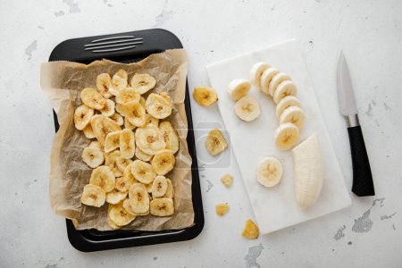 Foto de Bandeja para hornear con chips de plátano seco y plátano crudo con cuchillo de cocina.. - Imagen libre de derechos