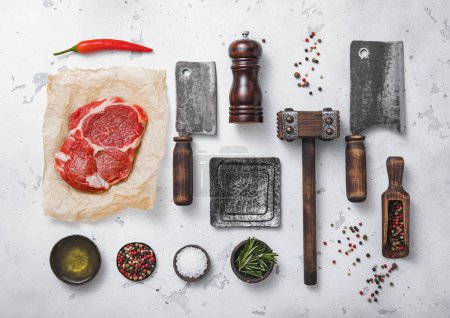 Photo for Red raw rib eye steak with meat cleavers,tenderizer,fork,knife and other kitchen meat utensils with oil,herbs,salt and pepper on light kitchen table background.Top view. - Royalty Free Image