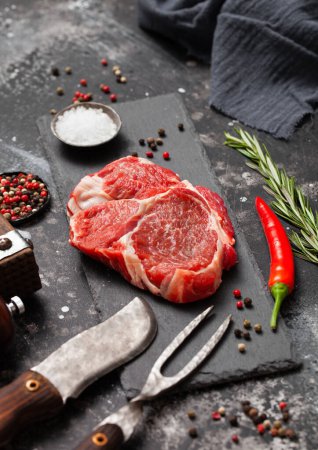Photo for Raw Rib eye fillet on stone board with fork and knife,salt and pepper with rosemary on black kitchen table. - Royalty Free Image