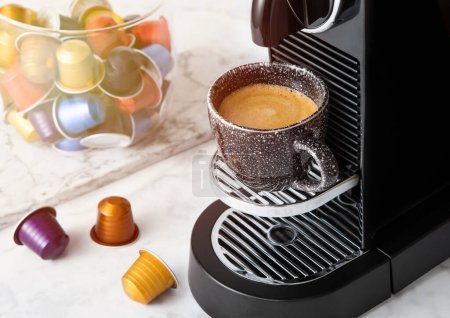 Foto de Taza de café negro y cafetera espresso casera con cápsulas de café con luz solar fondo de cocina. - Imagen libre de derechos