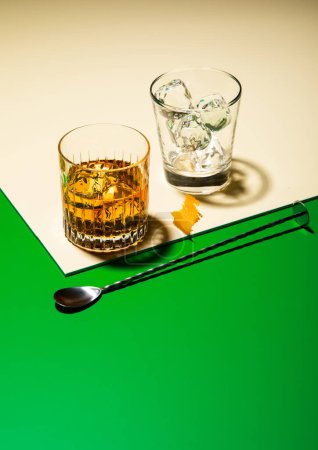 Photo for Glass of whiskey with ice and bar spoon with orange peel on green golden background.Concept art - Royalty Free Image