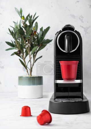 Téléchargez les photos : Tasse en acier expresso avec machine à café et dosettes rouges et olivier dans un fond de cuisine en marbre clair. - en image libre de droit