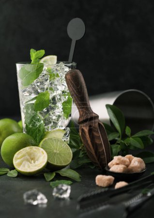 Photo for Glass of Mojito cocktail with ice cubes mint and lime on black board with spoon and fresh limes and cane sugar with wooden squeezer and steel shaker with ice.Best summer cocktail - Royalty Free Image