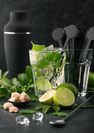 Photo for Glass of Mojito cocktail with ice cubes and disk stirrers,mint and lime on black board with spoon and fresh limes with cane sugar and black shaker. Best party drink. - Royalty Free Image