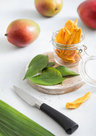 Foto de Mango seco en frasco de vidrio con frutas crudas y cuchillo sobre tabla ligera. - Imagen libre de derechos