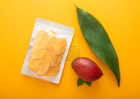 Téléchargez les photos : Grand paquet de tranches de mangue séchées avec des feuilles et des fruits crus sur fond jaune.. - en image libre de droit
