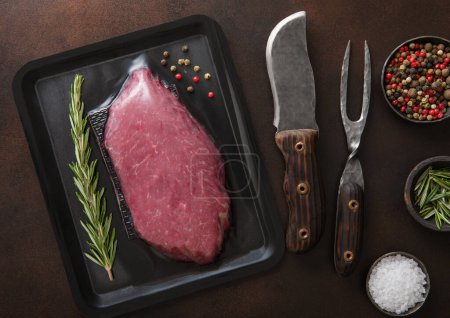 Photo for Beef raw fillet steak in vacuum tray with barbeque fork and knife on dark background with spices.Top view. - Royalty Free Image