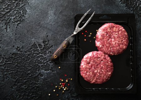 Photo for Raw minced beef burgers in plastic vacuum tray on black stone background with fork. Space for text - Royalty Free Image