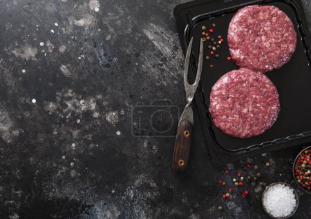 Photo for Raw beef burgers sealed in vacuum tray with fork and pepper on dark kitchen background.Top view. - Royalty Free Image