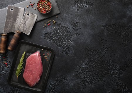 Téléchargez les photos : Bifteck de filet de boeuf cru dans un plateau à vide avec du poivre sur fond noir avec des coupe-viande. Vue du dessus - en image libre de droit