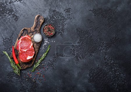 Foto de Tabla de cortar con pimienta y sal con romero sobre fondo negro y filete crudo de filete de costilla.. - Imagen libre de derechos