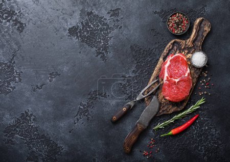 Photo for Raw rib eye steak fillet on wooden board with fork and knife,salt and pepper on dark kitchen table background.Top view. - Royalty Free Image