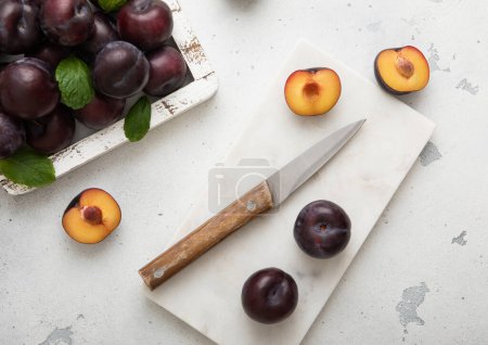Foto de Caja de madera de ciruelas moradas maduras crudas sobre fondo de cocina claro con cuchillo de cocina. - Imagen libre de derechos
