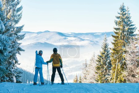 Foto de Pareja feliz en la estación de esquí. Hombre y mujer en los esquís tomados de la mano y mirando las montañas. Increíbles vacaciones de invierno - Imagen libre de derechos