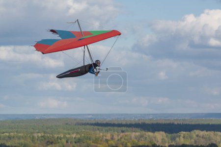 Photo for Colorful red hang glider wing soars over fields and forests with blue sky and clouds on the background. - Royalty Free Image