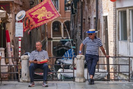 Téléchargez les photos : 2018-07-03 Venise, Italie. Gondoliers jeunes et vieux portant une tenue traditionnelle sur la jetée avec drapeau vénitien et canaux en arrière-plan - en image libre de droit