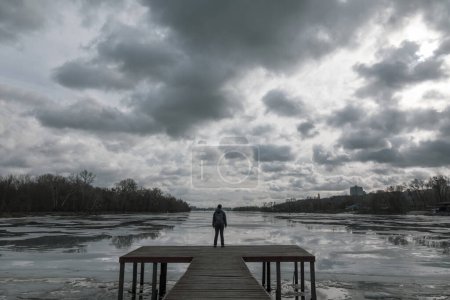 Téléchargez les photos : Femme solitaire debout sur la jetée en bois regardant les reflets des nuages dans l'eau froide. Photo dramatique sur les moments difficiles, les choix de vie difficiles et l'humeur dépressive - en image libre de droit