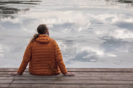 Téléchargez les photos : Homme solitaire en veste de randonnée orange assis sur la jetée en bois regardant le reflet du ciel dans l'eau - en image libre de droit