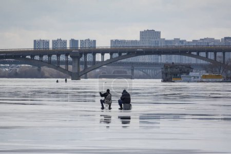 Téléchargez les photos : 2024-02-04 Kiev, Ukraine. Pêcheurs d'hiver sur la glace de la rivière Dnipro gelée avec des ponts et des immeubles de grande hauteur sur le fond - en image libre de droit