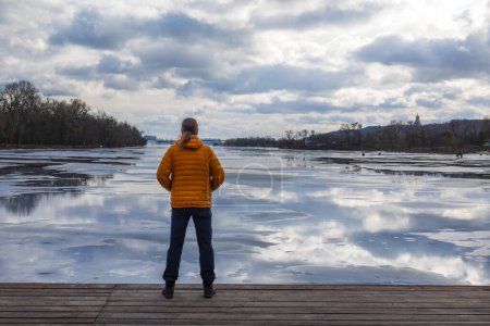 Téléchargez les photos : Homme solitaire en duvet orange debout sur une jetée en bois sur la rive de la rivière en regardant les reflets du ciel dans l'eau et la glace. - en image libre de droit