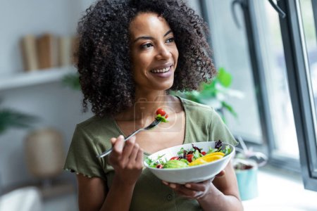 Photo for Shot of beautiful woman eating healthy salad while sitting on couch at home. - Royalty Free Image