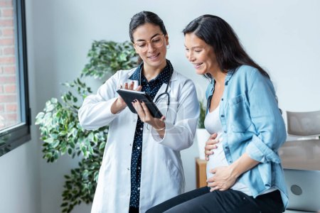 Téléchargez les photos : Prise de vue de gynécologue femme médecin montrant à la femme enceinte échographie bébé avec tablette numérique en consultation médicale. - en image libre de droit