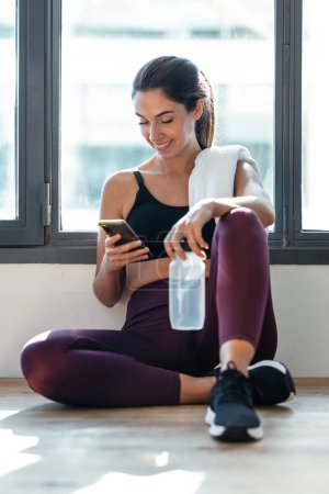 Téléchargez les photos : Prise de vue de femme sportive utilisant son téléphone portable tout en tenant une bouteille d'eau après un cours de pilates à la maison. - en image libre de droit