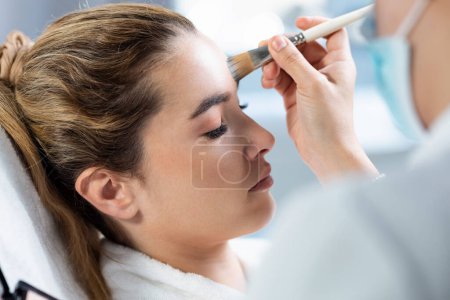 Téléchargez les photos : Prise de vue du cosmétologue appliquant le maquillage à une belle femme allongée sur une civière dans le centre de spa. - en image libre de droit