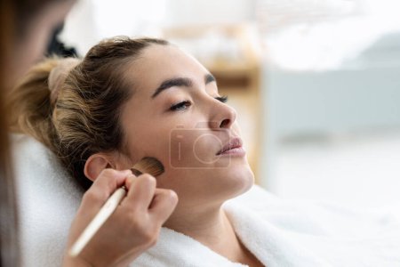 Foto de Foto de cosmetólogo aplicando maquillaje a una hermosa mujer acostada en una camilla en el centro de spa. - Imagen libre de derechos