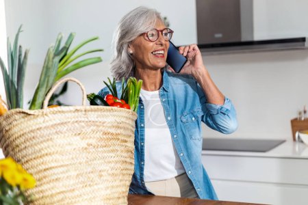 Photo for Shot of happy mature woman talking with smartphone in the kitchen at home. - Royalty Free Image
