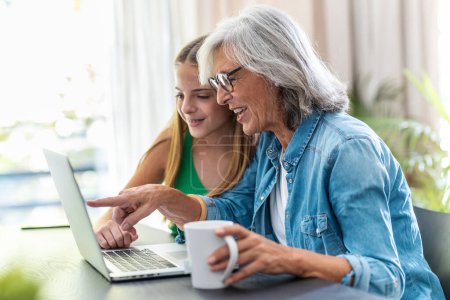 Photo for Shot of happy granmother shopping online with credit card using laptop with her granddaughter at home. - Royalty Free Image