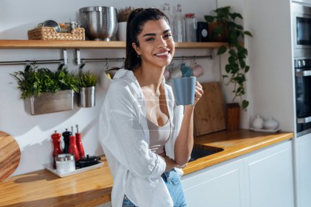 Foto de Shot of beautiful young woman drinking coffee while standing in the kitchen at morning - Imagen libre de derechos