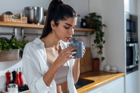 Téléchargez les photos : Shot of beautiful young woman drinking coffee while standing in the kitchen at morning - en image libre de droit