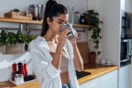 Téléchargez les photos : Shot of beautiful young woman drinking coffee while standing in the kitchen at morning - en image libre de droit