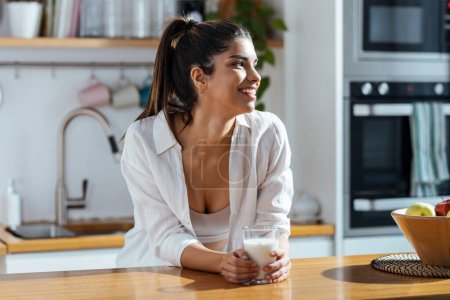 Téléchargez les photos : Tourné de belle femme boire un verre de lait tout en se tenant dans la cuisine le matin - en image libre de droit