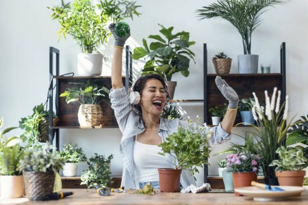 Téléchargez les photos : Shot of beautiful smiling woman arranging plants and flowers while dancing in a greenhouse - en image libre de droit