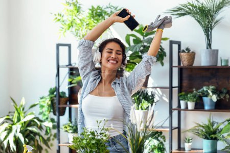 Téléchargez les photos : Shot of beautiful smiling woman arranging plants and flowers while dancing in a greenhouse - en image libre de droit