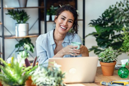 Foto de Shot of smiling woman drinking a cup of coffee while working with her laptop in greenhouse. - Imagen libre de derechos