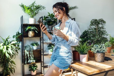 Téléchargez les photos : Shot of smiling woman drinking a cup of coffee while using her mobile phone in greenhouse. - en image libre de droit