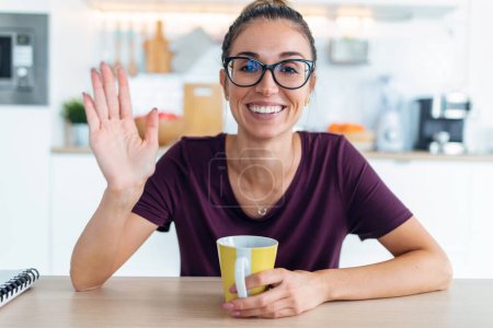 Photo for Shot of smiling young woman waving through the laptop web camera while holding a cup of coffee in the kitchen at home. - Royalty Free Image