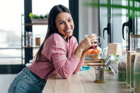 Téléchargez les photos : Tourné de belle jeune femme buvant du thé tout en regardant la caméra dans la cuisine le matin - en image libre de droit