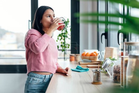 Téléchargez les photos : Tourné de belle jeune femme buvant du thé tout en se tenant dans la cuisine le matin - en image libre de droit