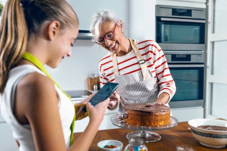 Téléchargez les photos : Tourné de mignonne petite-fille prenant des photos avec son smartphone tandis que grand-mère faisant gâteau au chocolat savoureux dans la cuisine. - en image libre de droit