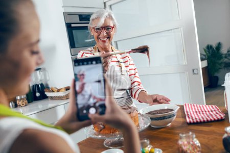 Foto de Shot of cute granddaughter taking photos with her smartphone while grandmother making tasty chocolate cake in the kitchen. - Imagen libre de derechos
