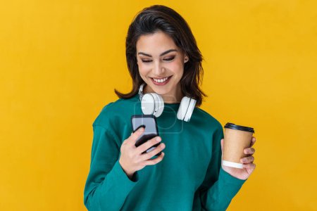 Téléchargez les photos : Portrait d'une femme rieuse utilisant un téléphone portable tout en tenant un café isolé sur jaune - en image libre de droit
